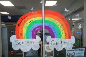 Artwork painted on the window of the Success Center at the Belleville Campus. It is a rainbow with the helping stations and kind words, saying "Help desk; Passion, Biology; Confidence, Math; Growth, Science; Optimism, English; Success, and Business; Wisdom, with "Success Center" written in the clouds under the rainbow.